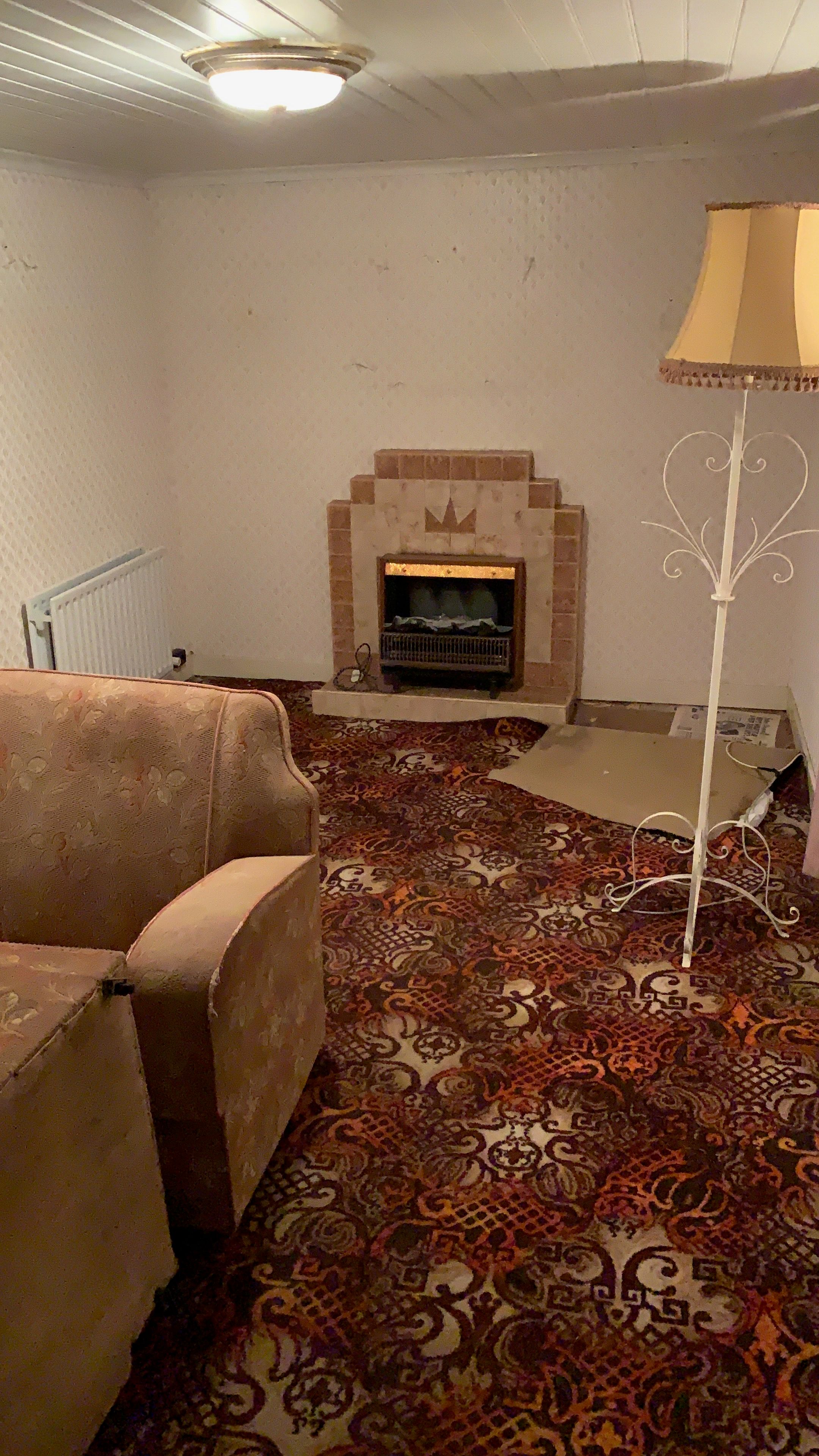 The old good room before renovation — patterned wallpaper, a tiled fireplace, a floral settee and a dense patterned carpet.