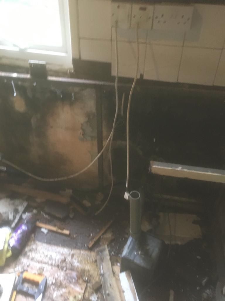 The old kitchen wall laid bare after the stainless steel sink and countertops were stripped out, pipework and wiring exposed.