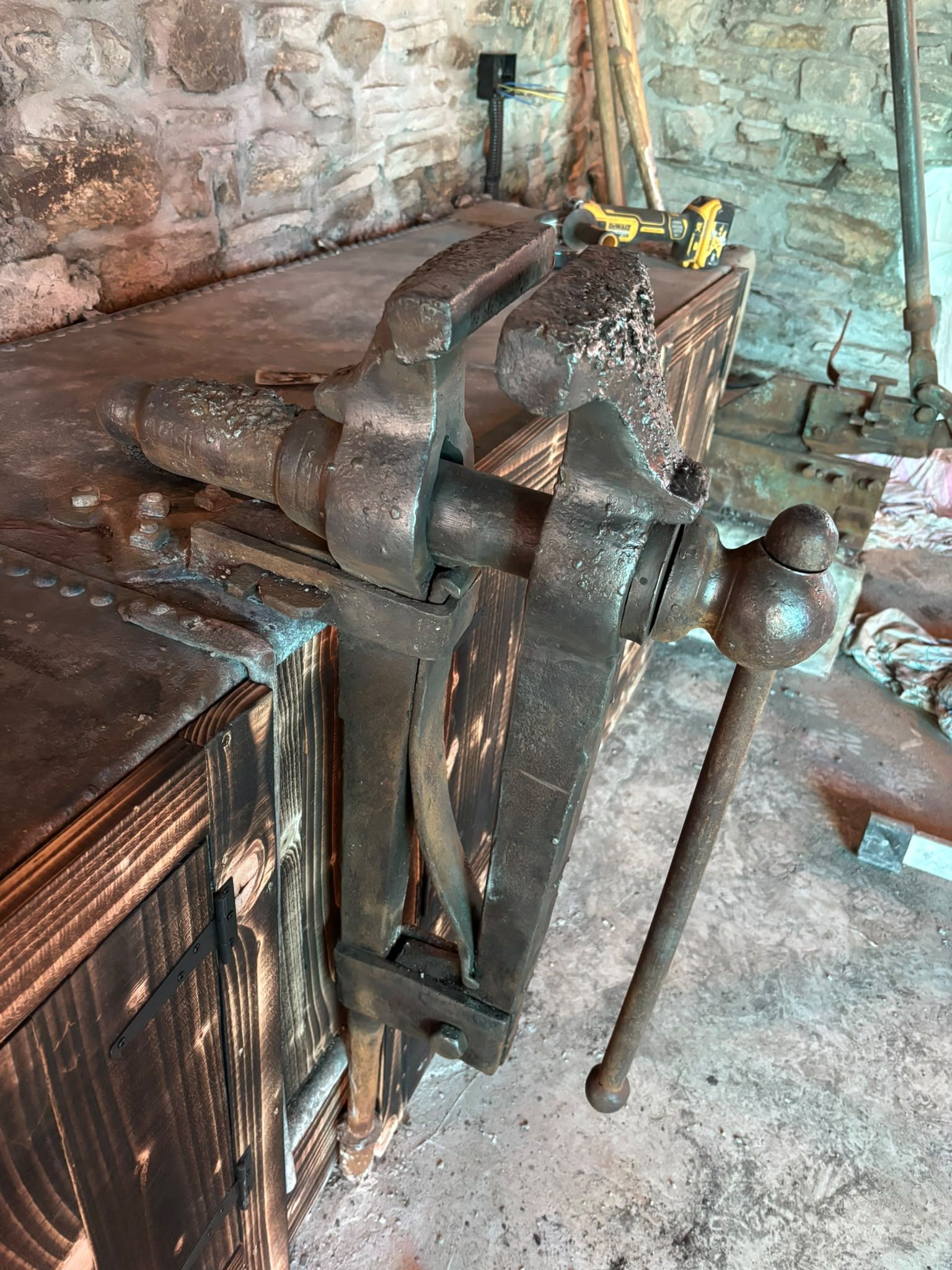 Davey John's large leg vice on a riveted metal bench with a charred timber front, against the old stone wall of the forge.