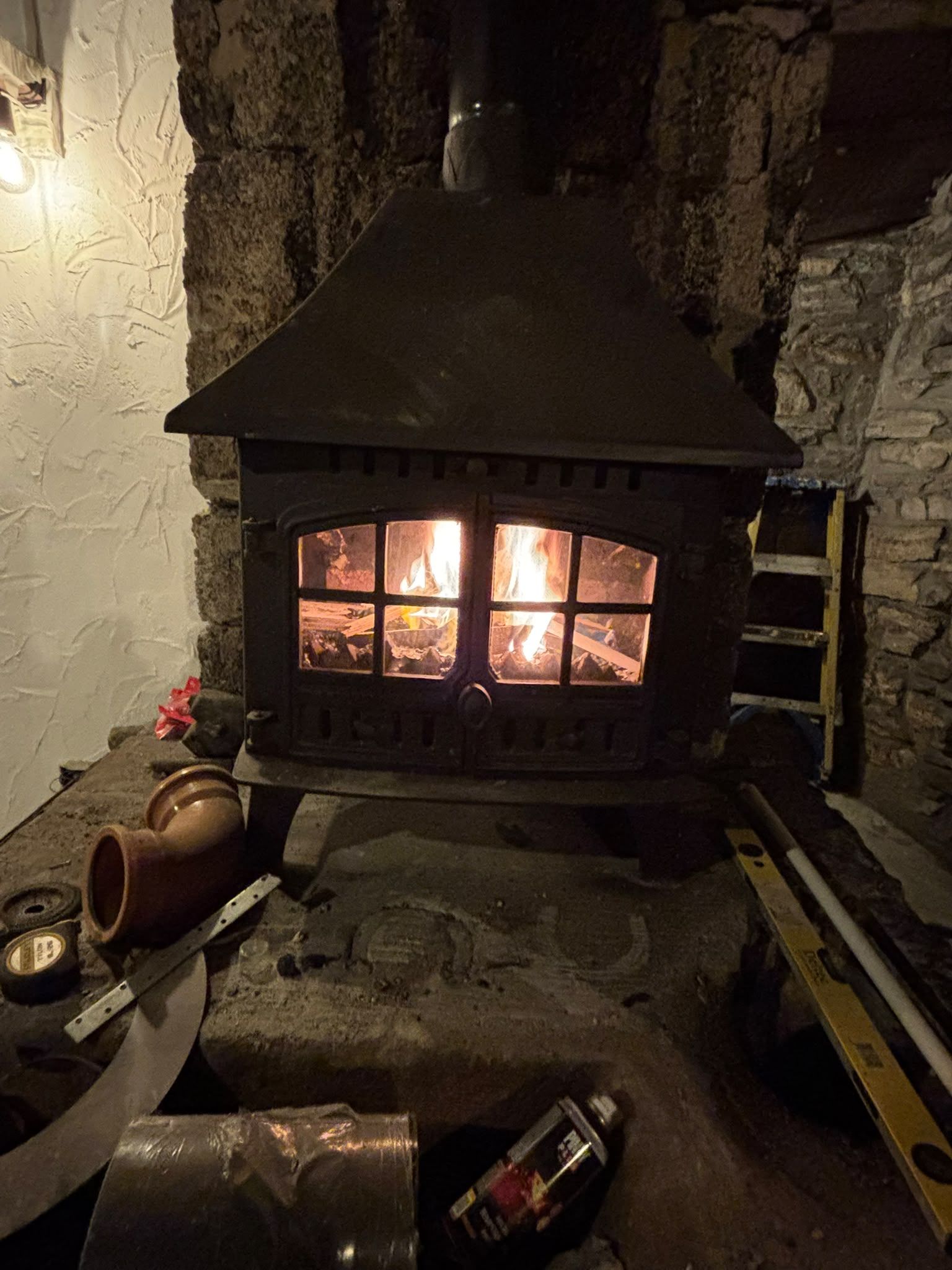The new wood stove lit in the old forge hearth, flames behind the glazed doors, tools and a spare pipe elbow on the stone hearth in front.