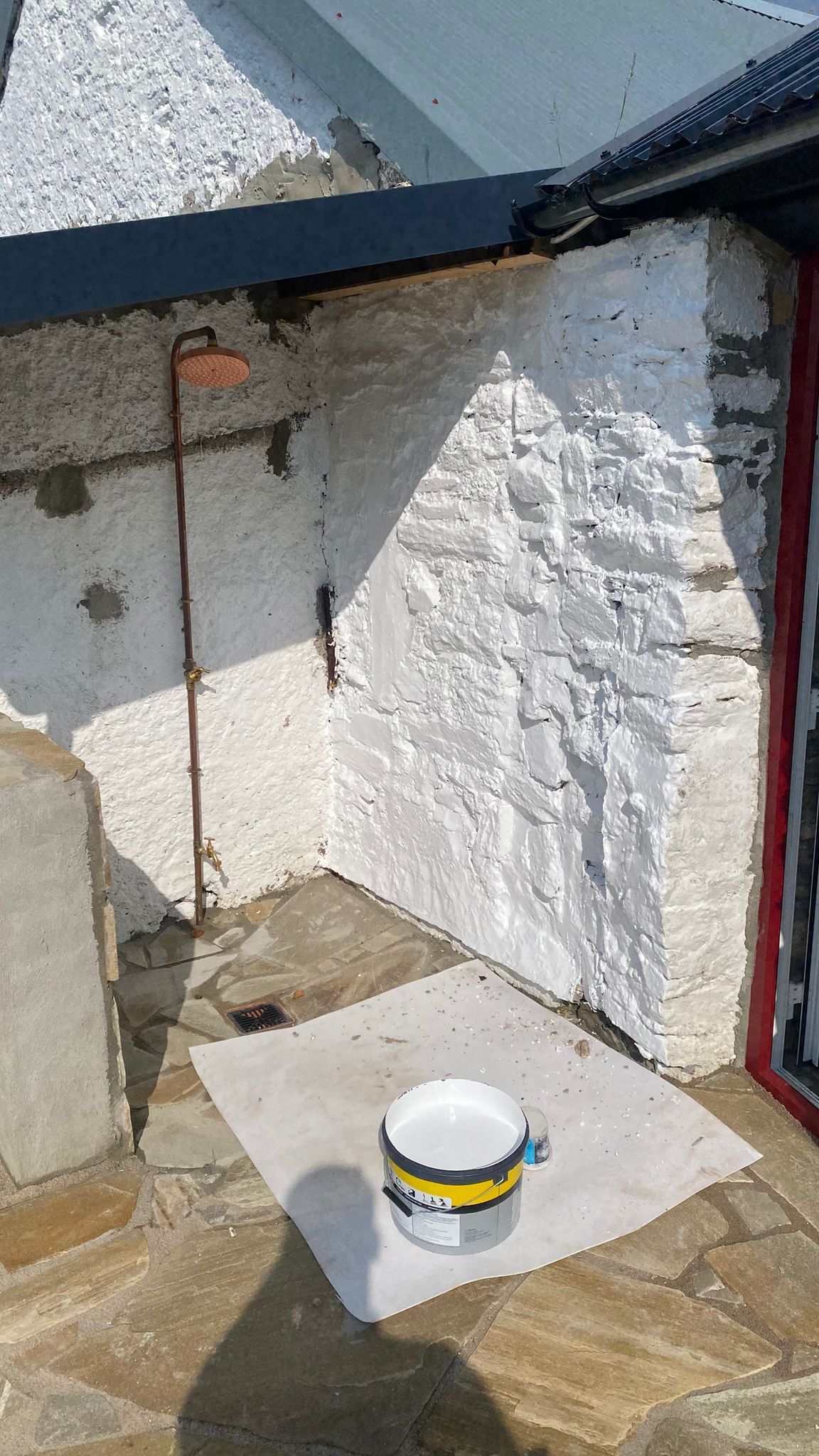The copper rainfall shower being fixed to the freshly whitewashed sauna wall, a drain set into the stone-flag paving.