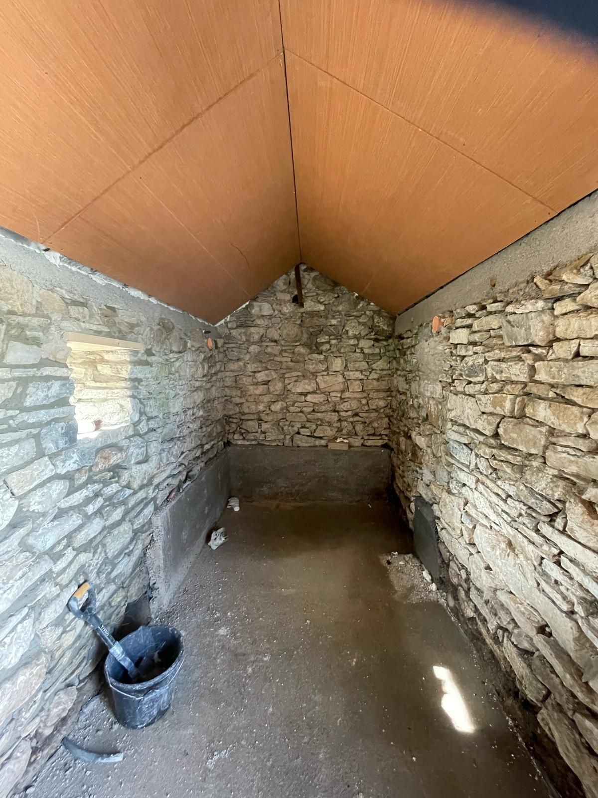 The sauna shell closed in — poured concrete floor, a pitched ceiling over the dry-stone walls, the left wall freshly pointed.