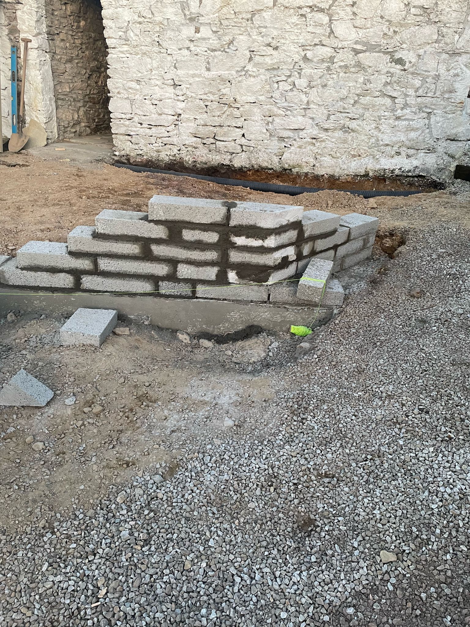 A new blockwork wall being built at the base of the whitewashed sauna wall — the start of the retaining wall that holds the level terrace at the door.