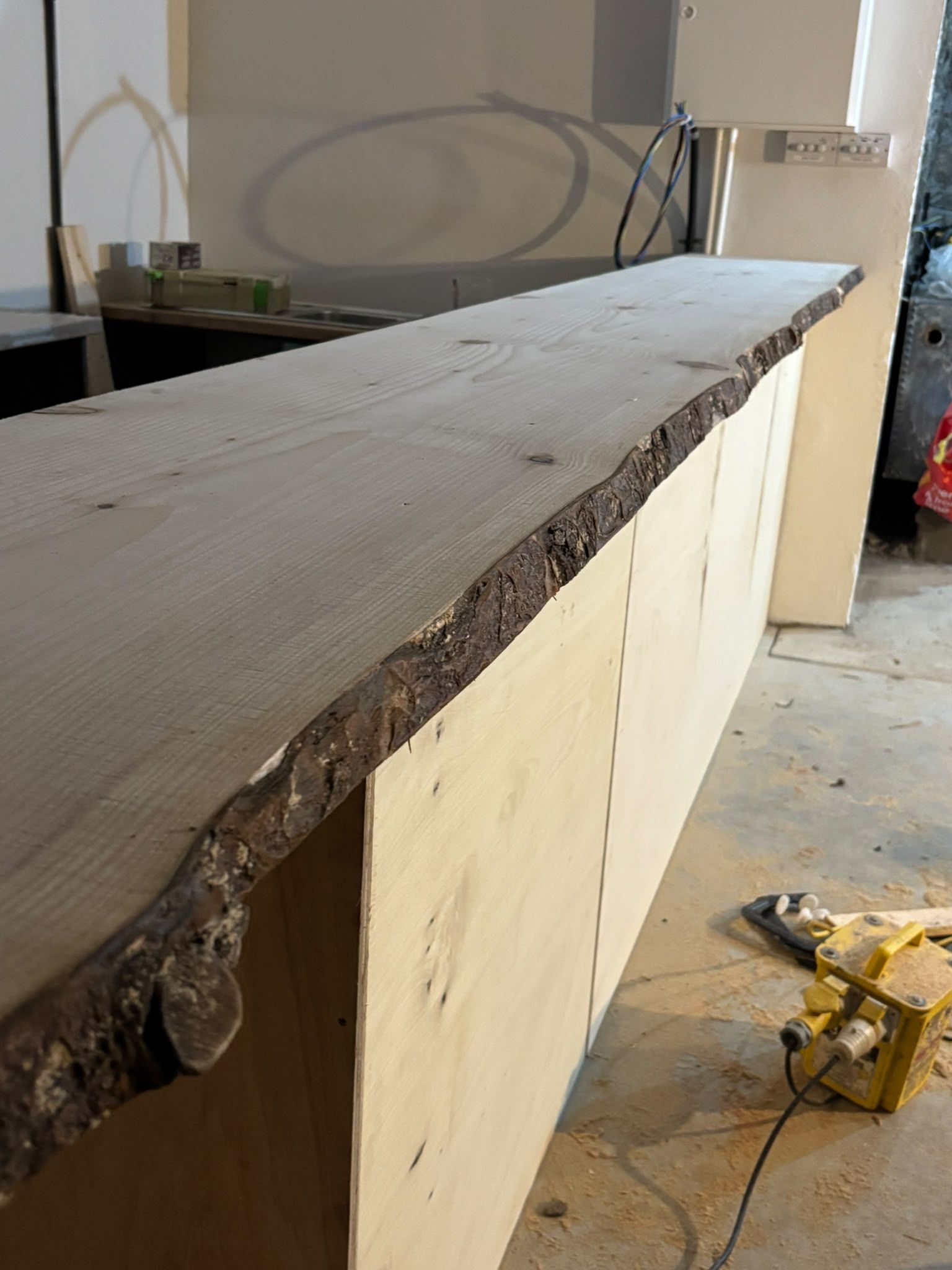 The bar carcass in bare plywood with a live-edge pine top laid on it, the bark still on the wavy front edge, sawdust and a yellow site power box on the floor.