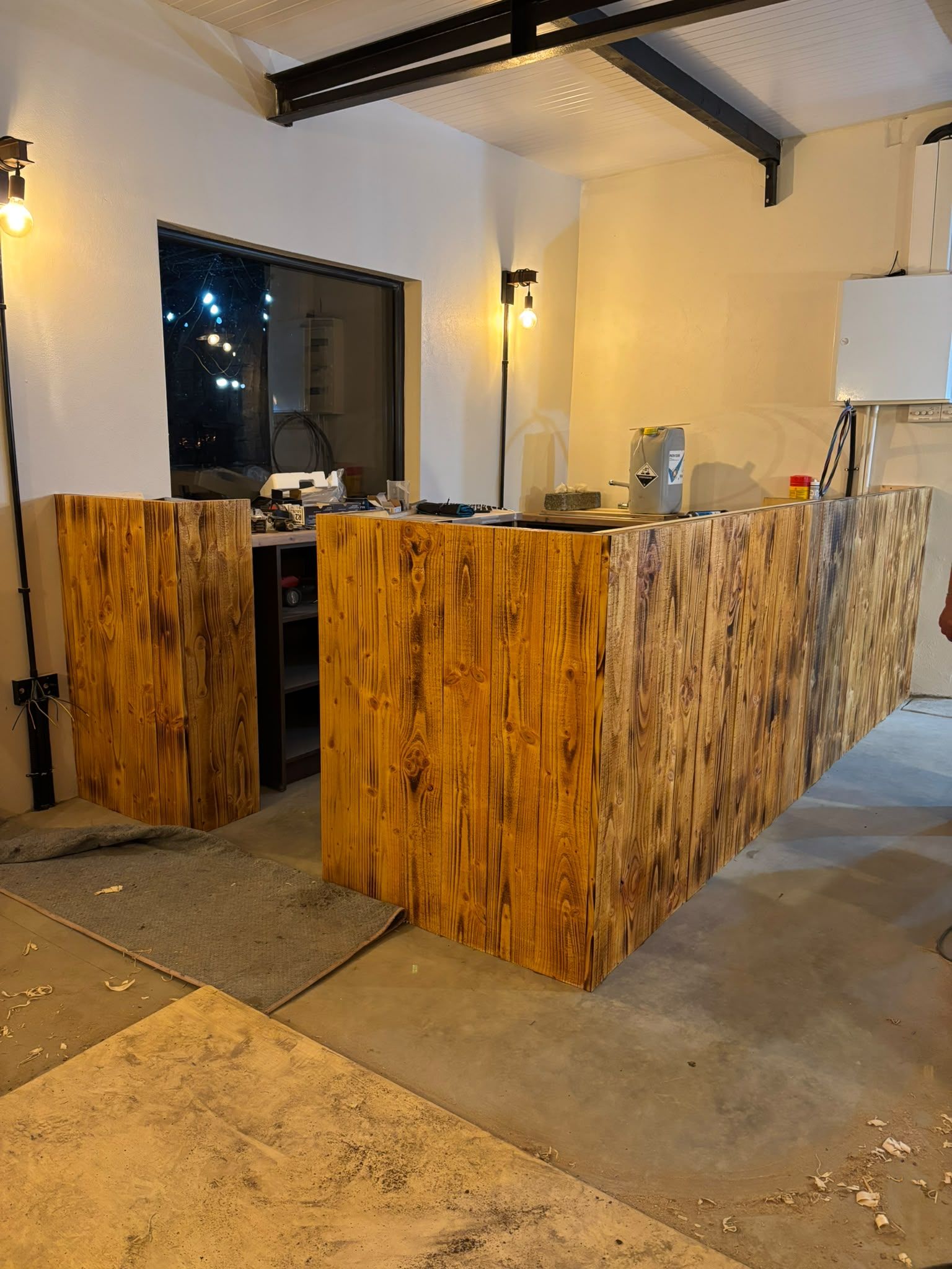 The L-shaped bar clad in scorched timber at night, wall lights lit, black steel trusses overhead and a dark window reflecting string lights.