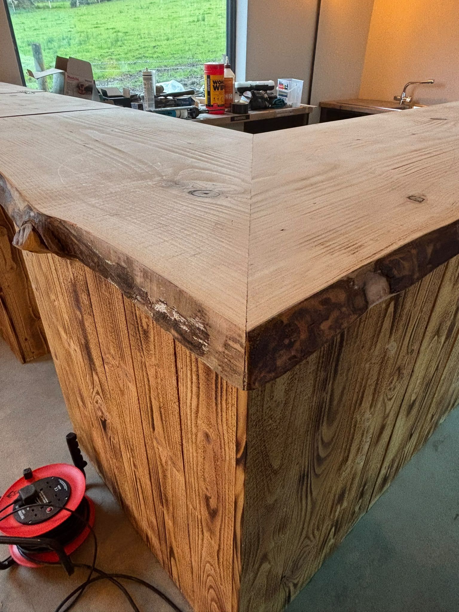 The corner of the bar where two live-edge pine planks meet on a mitre, scorched-timber cladding below and a green field through the window behind.