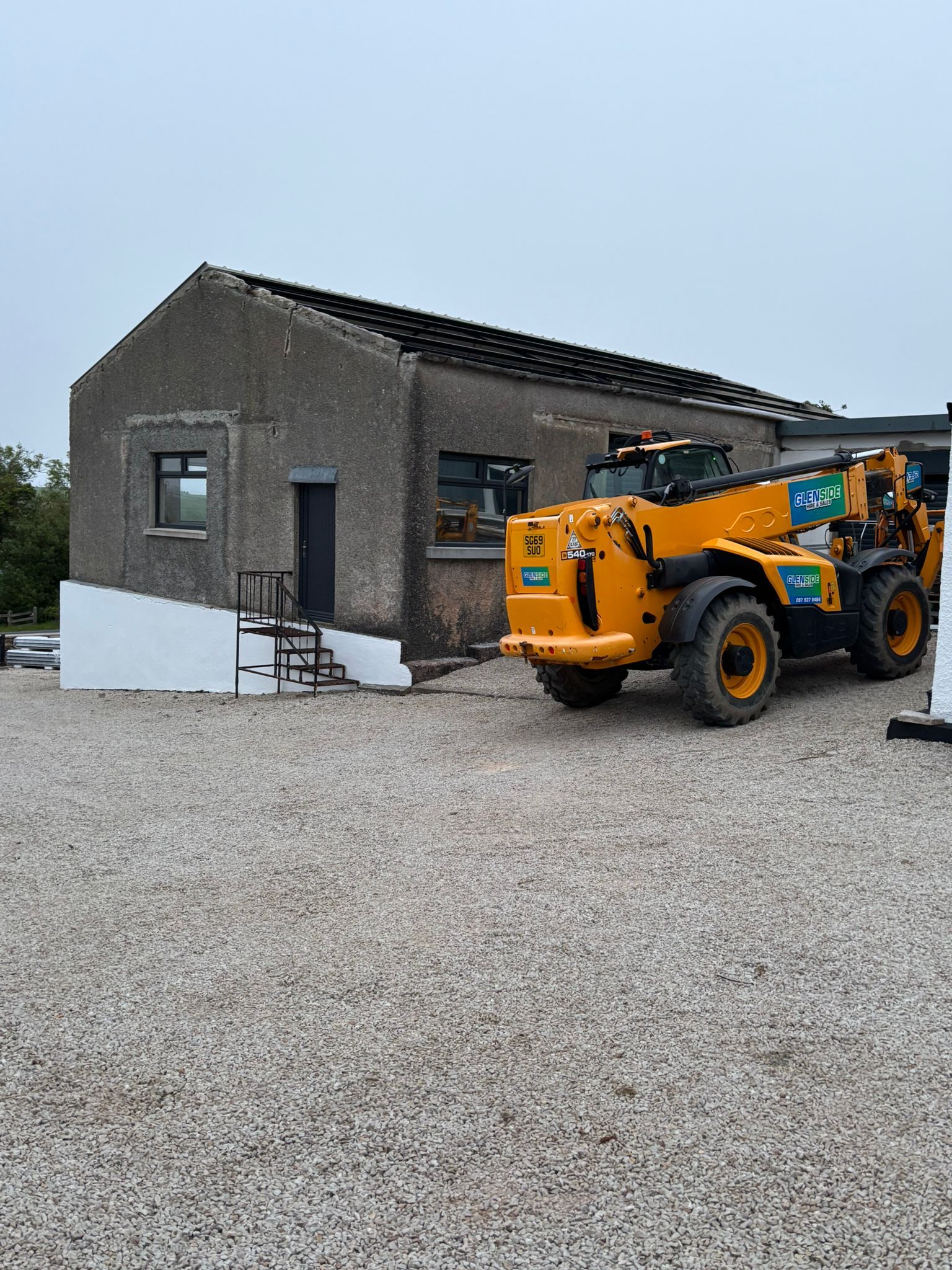 The workshop from the gravel yard, the old covering coming off the roof, a telehandler alongside.