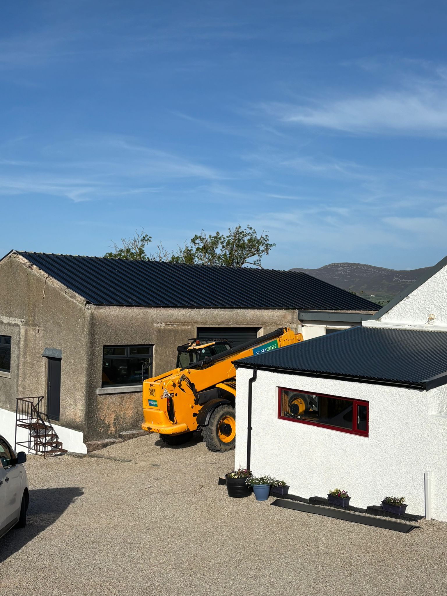 The workshop with its new dark roof in bright sun, a telehandler at the roller door, the white games room with its red window alongside.