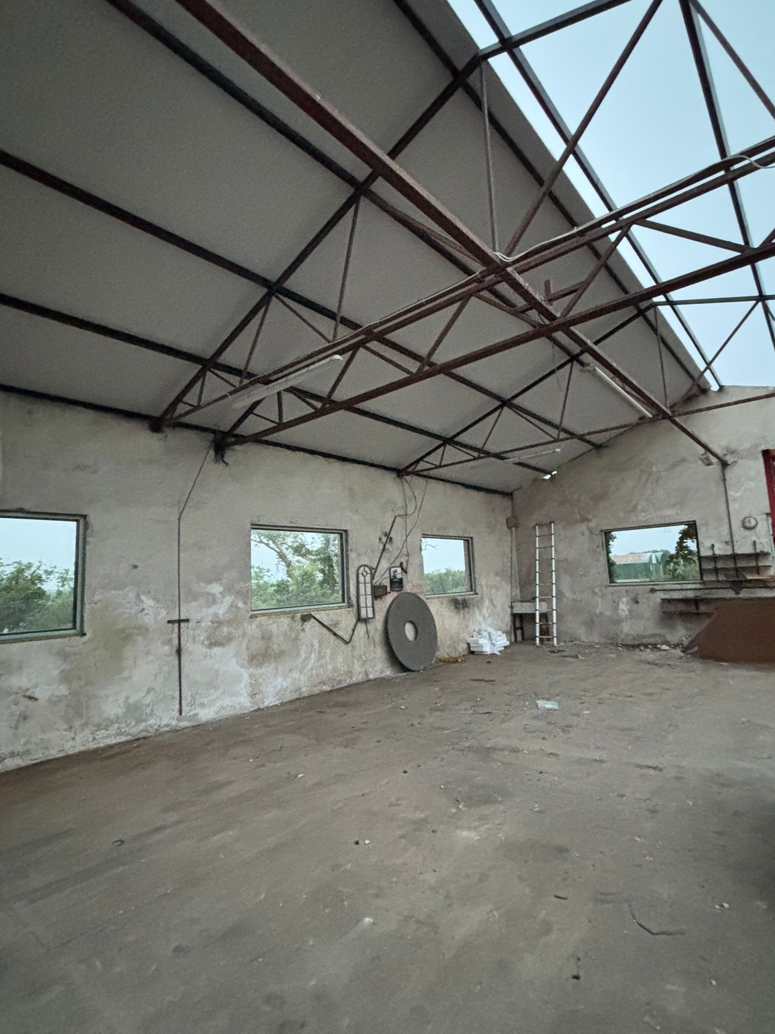 Inside the stripped workshop — the steel roof trusses overhead, an old circular grindstone leaning against the bare render wall, windows to the fields.