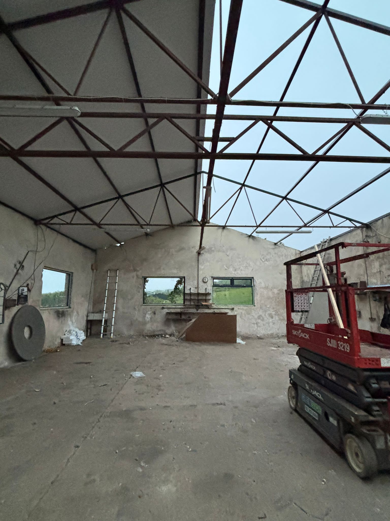 The bare workshop interior stripped back, exposed steel trusses overhead, a ladder against the weathered render and the windows looking out to green fields.