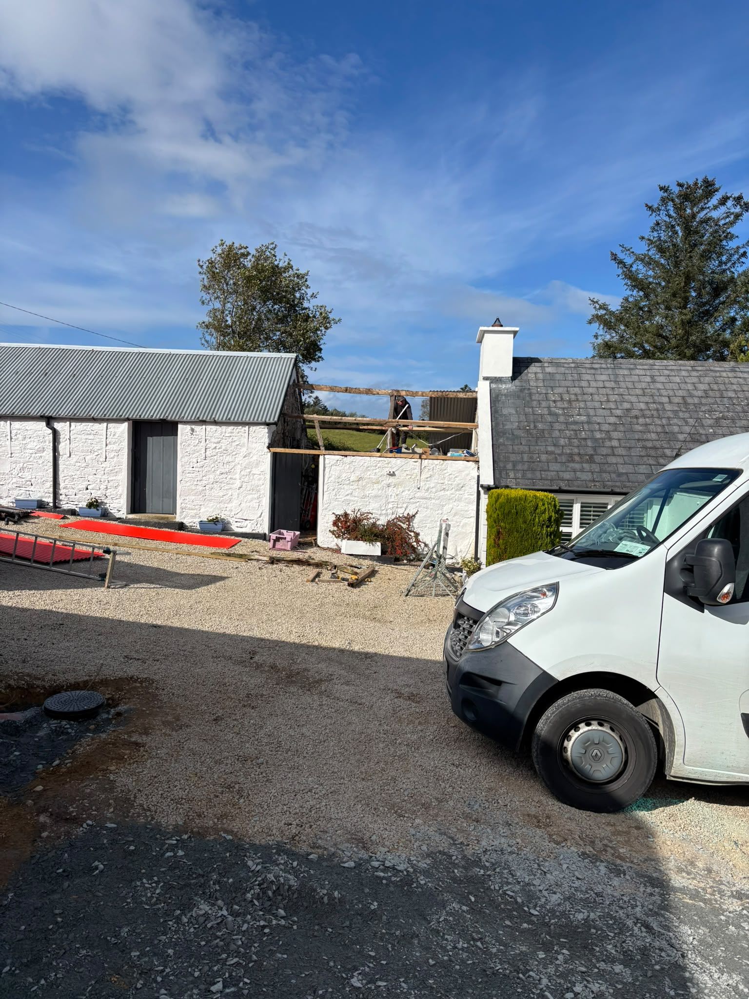 The low outbuilding between a whitewashed shed and the cottage with its roof stripped to the rafters under a blue sky, red corrugated sheets lying on the gravel.