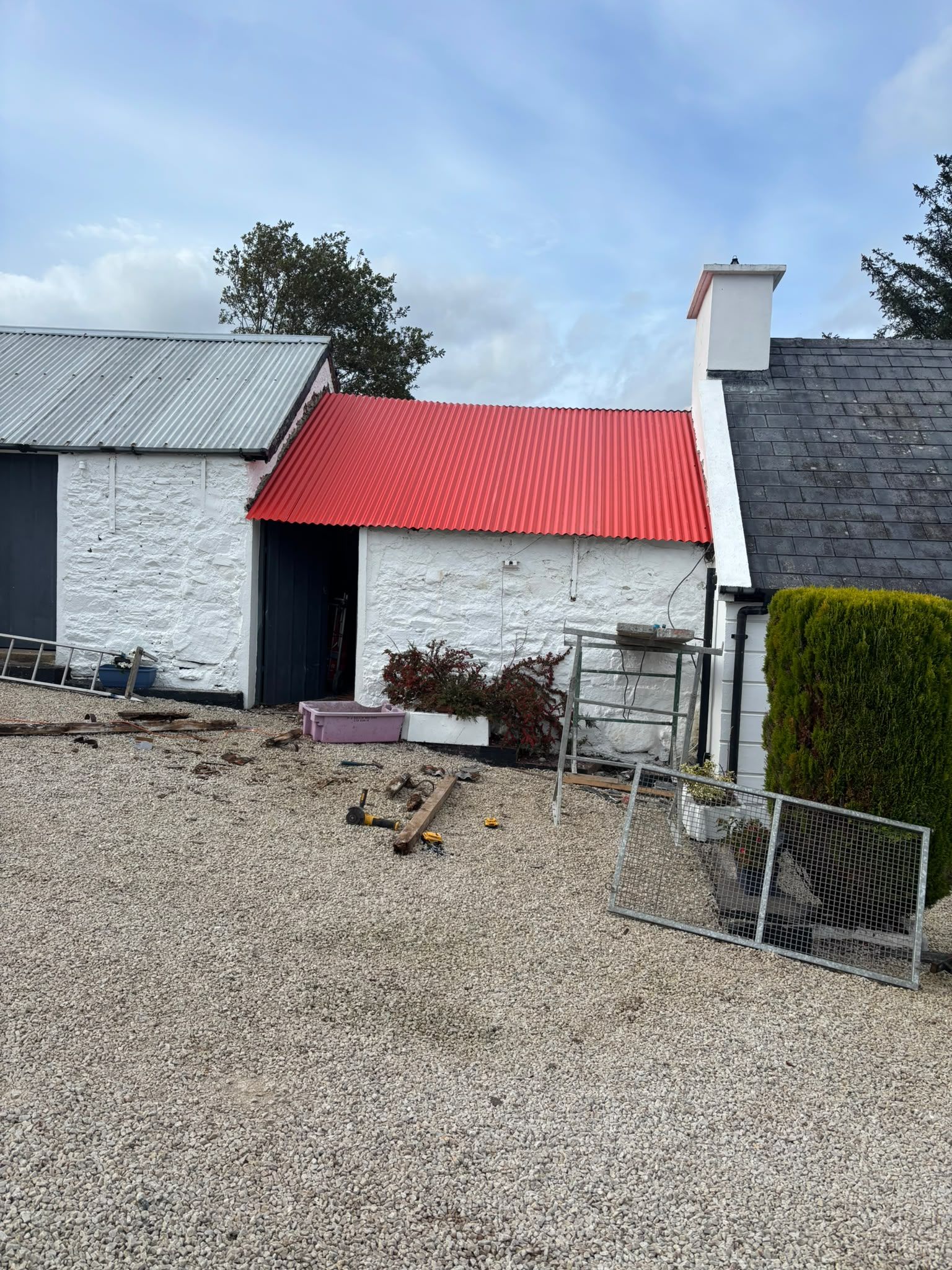 The same outbuilding with a new red corrugated roof on, white walls and dark doors beside it and the cottage's slate roof and chimney to the right.