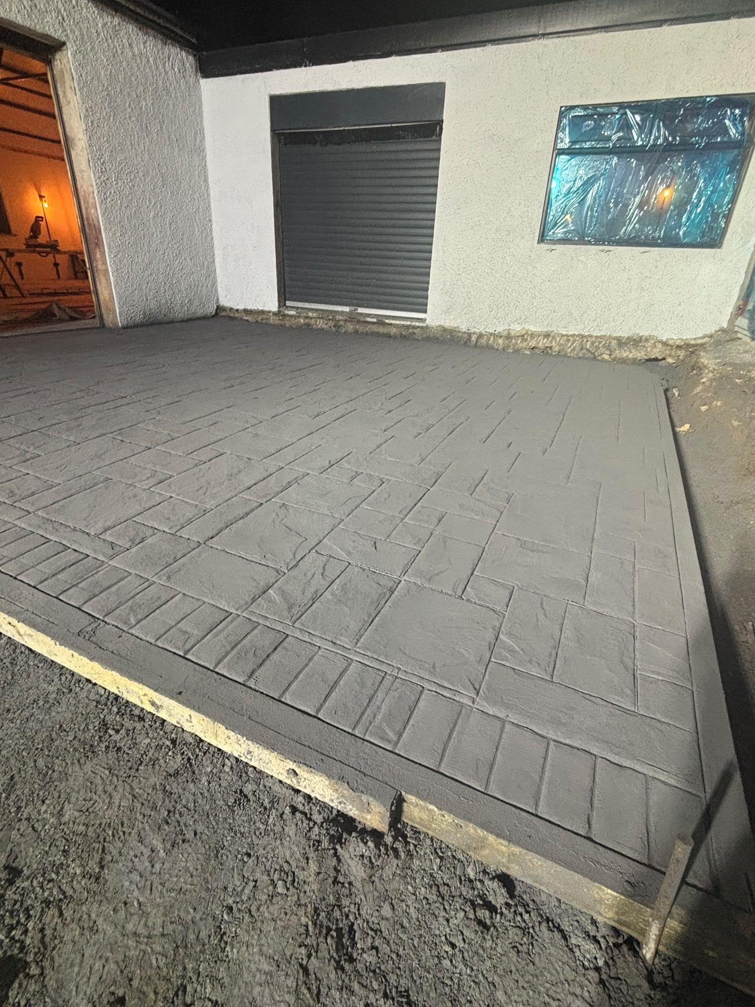 The stamped slab at night seen at an angle, showing a cobble-pattern border around a flagstone-pattern field, the forge doorway glowing orange at the left.