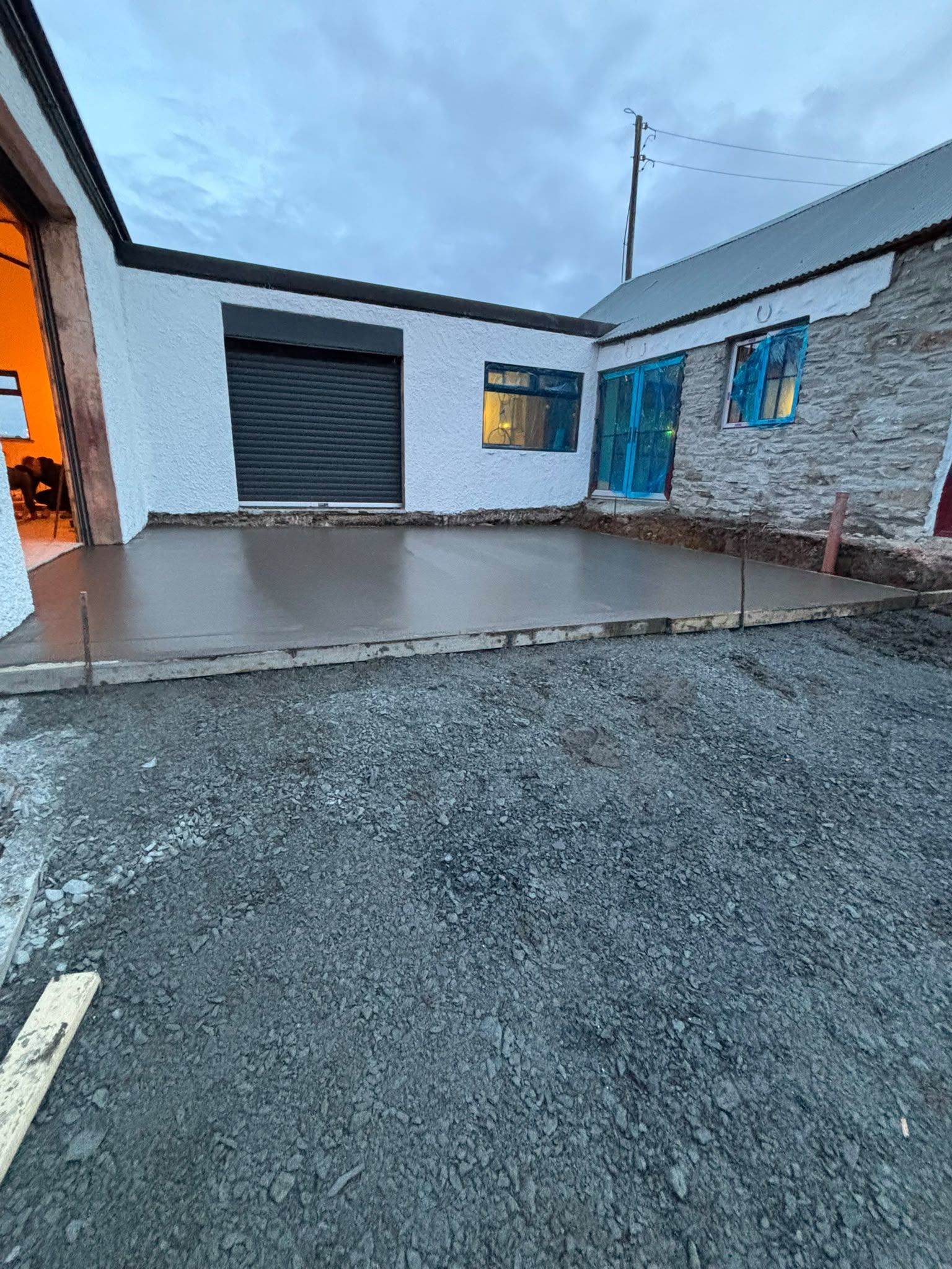 The freshly poured and floated concrete slab at dusk in the courtyard, timber shuttering along its edge, white rendered walls, a dark roller shutter and new windows in blue film, the forge doorway glowing orange at the left.