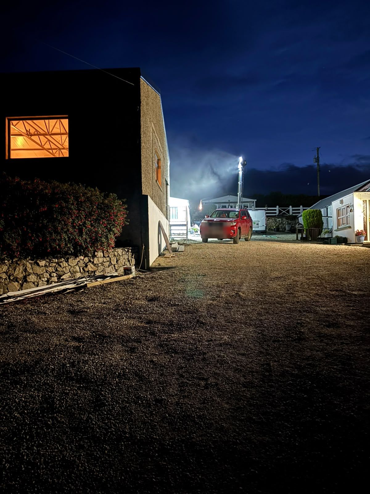 The forge at blue hour, a lit window showing the roof trusses, a tall floodlight tower at the end of the gravel yard and the white cottage to the right.