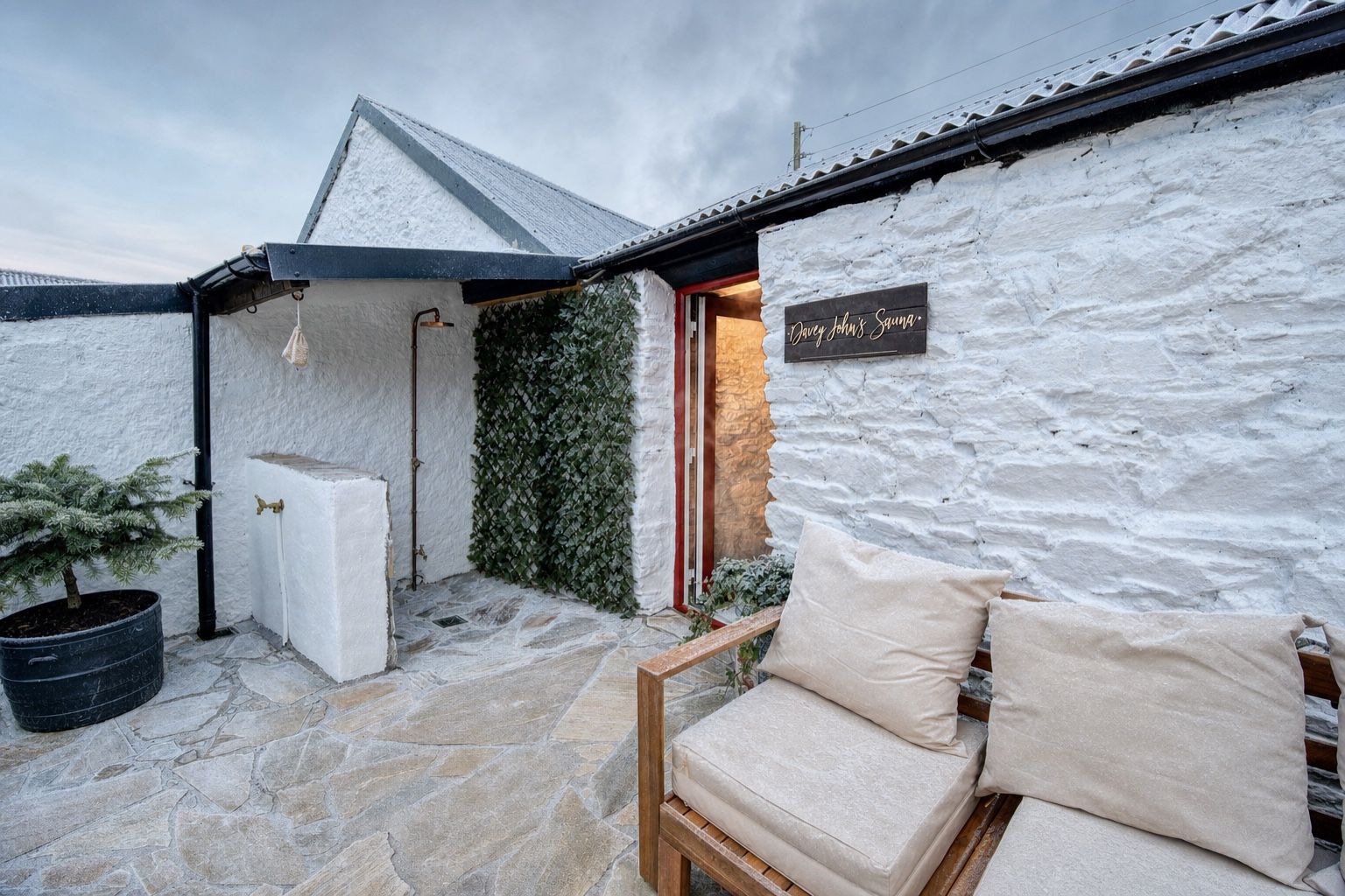 The sauna door open on a frosty morning, warm light spilling onto the frost-silvered crazy-paving patio, the copper shower and teak sofa either side